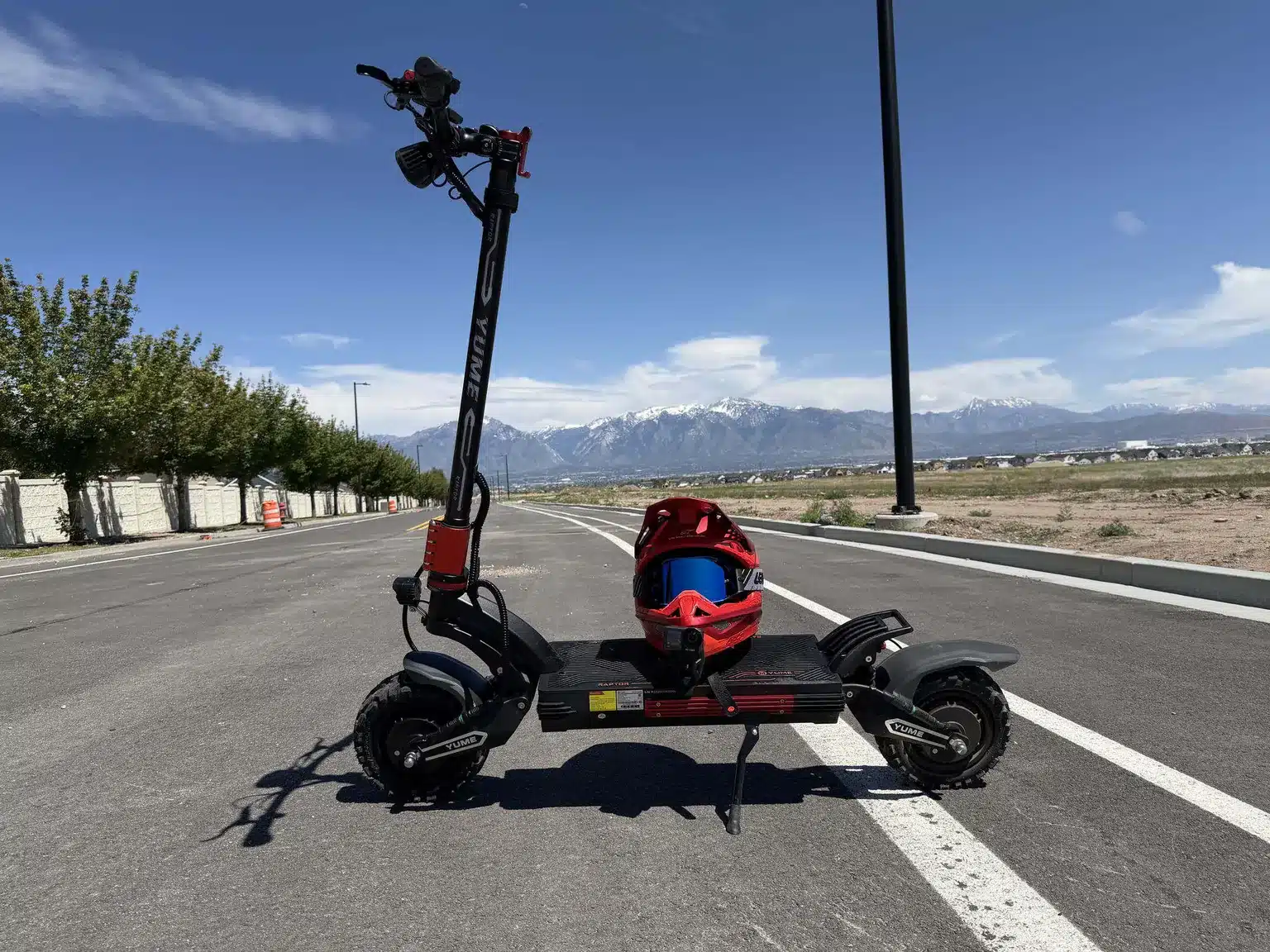 YUME Raptor 2 Review: Fastest Electric Scooter Under $1,600 1 YUME Raptor 2 electric scooter parked on a road with a red full-face helmet on the deck and snow-capped mountains behind